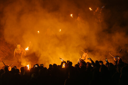 Crowd watches figures in horned Krampus costumes holding torches amidst smoke and fire. Krampus and St Nicholas, 5 December in Camporosso Valcanale, Tarvisio, Italy.