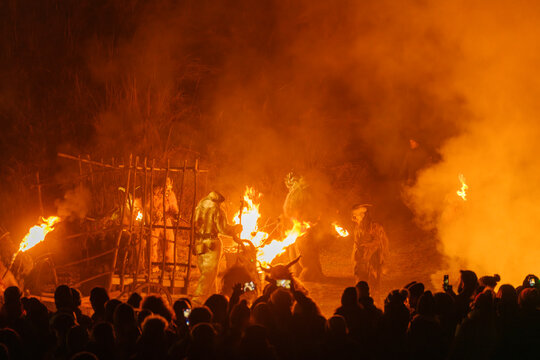 Crowd watches figures in fur costumes and horned masks holding torches amidst smoke and fire. Krampus and St Nicholas, 5 December in Camporosso Valcanale, Tarvisio, Italy.