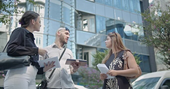 Three young business people stand outside an office building discussing something on a tablet and printed papers on a sunny day.