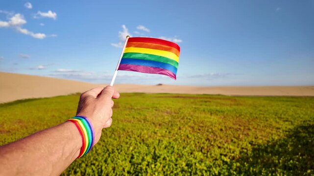 Slow motion video of a tourist waving a small rainbow flag in Maspalomas, Gran Canaria, with sand dunes and green vegetation in the background