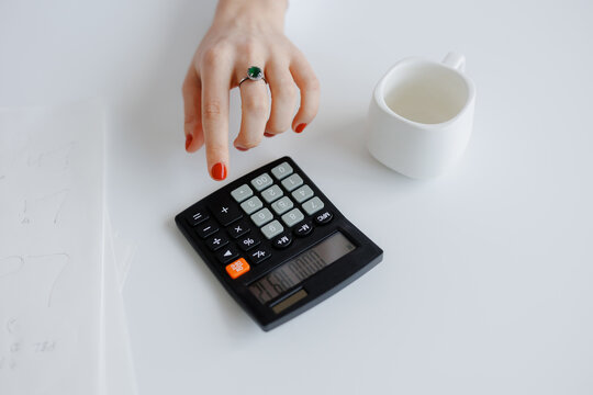 Hand pressing calculator buttons with empty cup on white table during work session