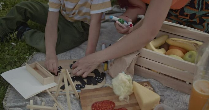 Mother and son enjoy a picnic in the park, playing dominoes and drawing together.