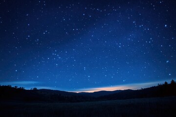 Clear night sky filled with numerous bright stars over a dark silhouetted landscape of rolling hills and distant trees at twilight