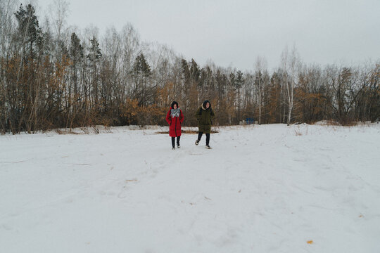 Two individuals stroll across a vast expanse of white snow amidst a nearly bare forest.