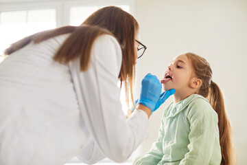 Female doctor pediatrician laryngologist with tongue depressor asks little child girl to open mouth...