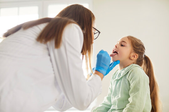 Female doctor pediatrician laryngologist with tongue depressor asks little child girl to open mouth for throat, larynx and pharynx diagnostic examination for strep bacteria, flu, or pharyngitis
