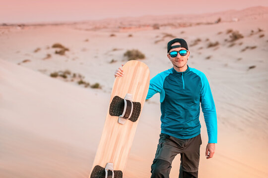 Male athlete holding sandboard, walking on sand dune, getting ready for sandboarding in desert at sunset