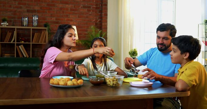 Family eating panipuri snacks together at home, Indian father, mother, son daughter enjoying golgappa at dining table in cozy living room while bonding, chatting, laughing and spending quality time