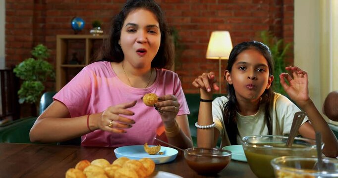 Family eating panipuri snacks together at home, Indian father, mother, son daughter enjoying golgappa at dining table in cozy living room while bonding, chatting, laughing and spending quality time