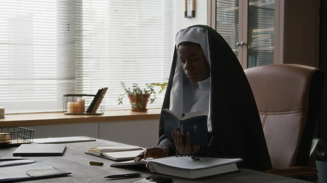 Black young religious sister dressed in habit attire reading bible book and taking notes while sitting in leather chair at large desk in office
