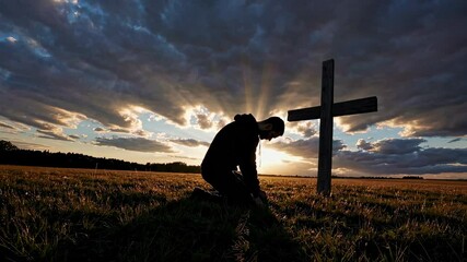 Man kneeling in prayer by cross at sunset on grassy field with dramatic clouds and radiant light, spiritual faith and repentance concept