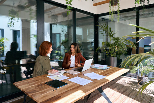Professional ladies employee and manager having conversation using laptop at work. Two busy business women workers team of young and middle age talking in creative green office sitting at desk.