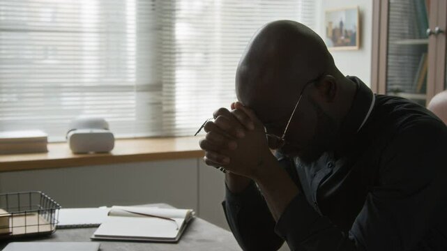 Tilt up view of bald African American young priest dressed in black clerical shirt sitting at desk with his hands clasped in prayer and head bowed