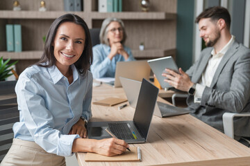 Attractive Caucasian young confident businesswoman sitting at the office table with group of colleagues in the background, working on laptop computer making notes looking at camera