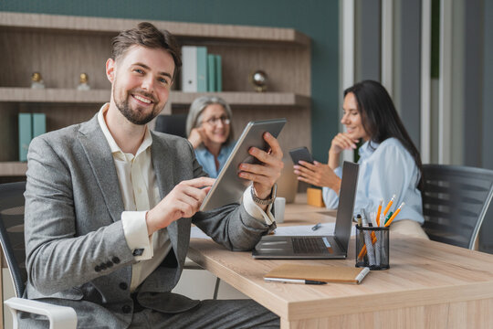 Smiling Caucasian young confident businessman sitting at the office table with group of colleagues in the background, working holding using digital tablet laptop computer looking at camera - Powered by Adobe