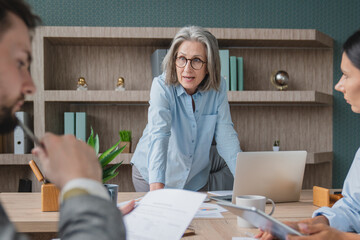 Senior older female executive ceo and happy multicultural business people discuss corporate project at boardroom table. Smiling diverse corporate team working together in modern meeting room office.