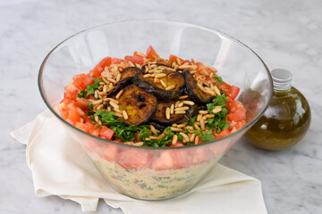 Mediterranean Couscous Salad with Grilled Eggplant on napkin featuring fluffy couscous, tomatoes, parsley, grilled eggplant slices, and toasted pine nuts with a olive oil bottle side view