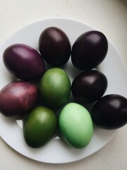 Flat lay of colorful dyed Easter eggs in green and purple shades on a white plate with soft indoor light perfect for spring holiday decor, family traditions and creative food styling visuals