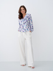 Smiling woman with long brown hair wears a colorful floral blouse and white wide-legged trousers,...