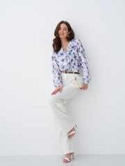 Elegant woman in floral blouse and white trousers posing against a plain background
