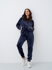 Smiling woman in dark casual tracksuit with hood, hands in hoodie pocket, stands against light wall. She wears white sneakers and has wavy hair, creating a relaxed, stylish look