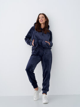 Smiling woman in dark casual tracksuit with hood, hands in hoodie pocket, stands against light wall. She wears white sneakers and has wavy hair, creating a relaxed, stylish look