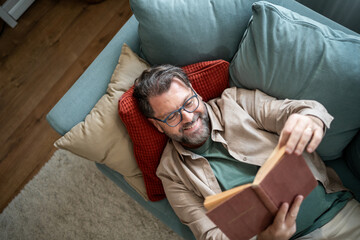 Fototapeta premium Smiling man lying on sofa reading a book at home