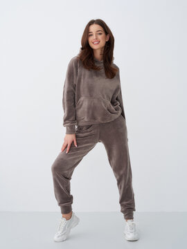 Young woman wearing a cozy, brown velour tracksuit looking confident and smiling, standing in a relaxed position, white sneakers, long brown hair, neutral light gray backdrop
