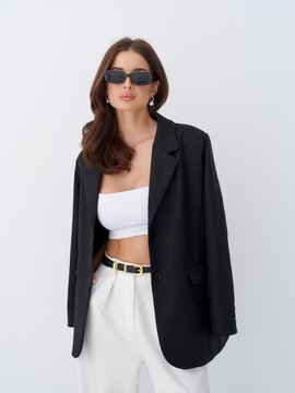Woman with long brown hair wears oversized black blazer and sunglasses, paired with a white tube top and high-waisted pants. She exudes a chic and fashionable look