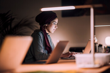 A Moroccan Arab Muslim woman is working intently on her laptop at her desk. She is focused on typing in a modern office environment at night.