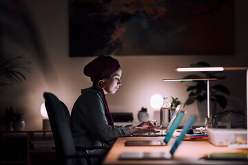A dedicated young Moroccan Arab Muslim woman works on her laptop in her home office at night. She is focused on her tasks in the quiet space.