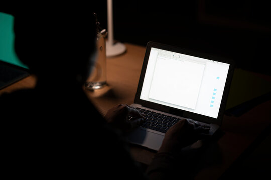 A Moroccan Arab Muslim person works on a laptop at night. Typing on the keyboard, illuminated by the screen, focusing on their task in the dark.