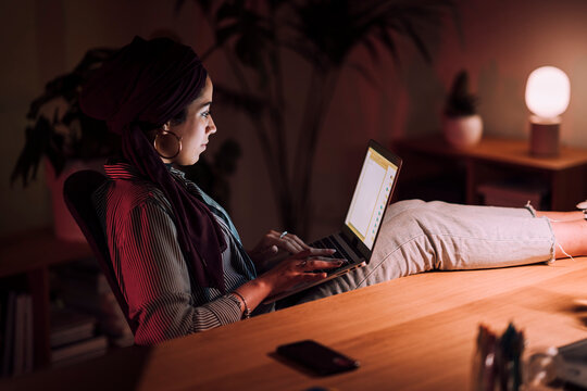 A young Moroccan Arab Muslim woman in a hijab works on her laptop at a desk in a relaxed pose with her feet up. She is focused on her work in the evening light.