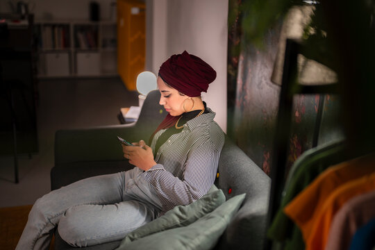 A stylish Moroccan Muslim woman wearing a hijab relaxes on a sofa. She is using her smartphone, possibly texting or browsing social media at home. - Powered by Adobe
