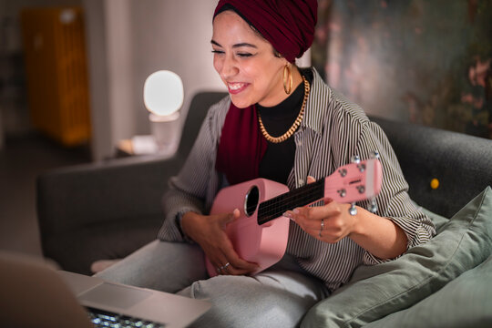 A smiling Moroccan Muslim woman wearing a hijab learns ukulele online. She is sitting on a sofa with a laptop, enjoying her musical hobby at home.