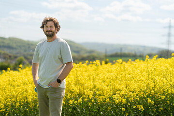 Fototapeta premium Cute bearded curly man in T-shirt smiling standing in front of yellow blooming rapeseed field. Concept of emotions, leisure and travel