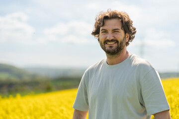 Cute bearded curly man in T-shirt smiling standing in front of yellow blooming rapeseed field. Concept of emotions, leisure, nature and travel