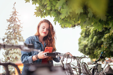 Portrait of smiling red-curled long-haired Caucasian teenage girl locking bike at bicycle-sharing station while using modern smartphone. Green urban transport and modern technology devices concept