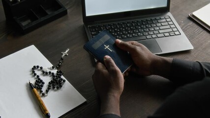 Over the shoulder view of unrecognisable Black priest in clerical shirt holding bible book with both hands while sitting at wooden table with laptop during workday