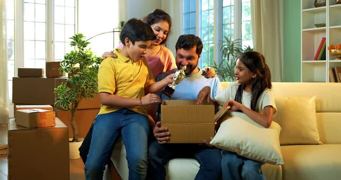 Indian family unboxing at new home, Asian young father, mother two kids eagerly taking out belongings from box while sitting on sofa together in their lavish new residence with smiles