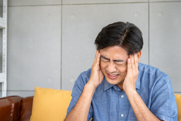 Asian elderly man suffering from intense headache or migraine, holding head in pain while sitting on sofa
