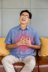 Asian elderly man experiencing chest pain, possible heart attack, sitting on a brown leather sofa