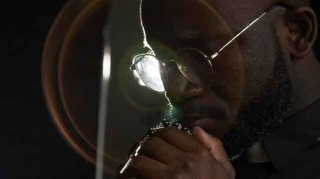Close-up view of Black young catholic priest wearing round glasses in traditional clerical shirt with tonsure collar praying to god with hands put to his chin