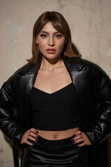 A young woman stands confidently in a black leather jacket and crop top, exuding a modern...