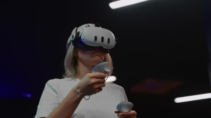 Young woman in VR headset with joysticks, fully immersed in virtual space while adjusting game or program settings. Captures modern interactive technology experience.