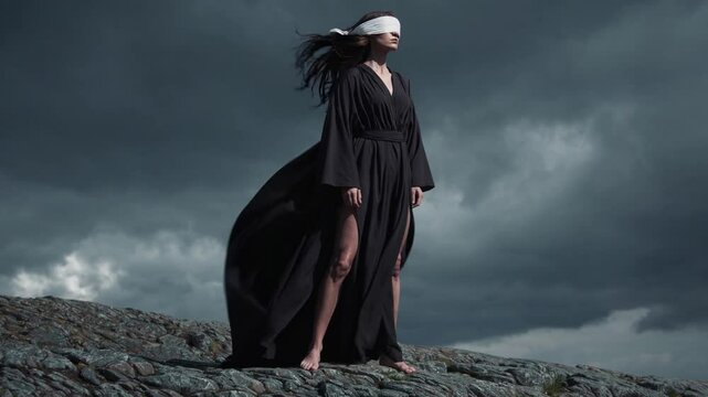 Femme en robe noire et les yeux band&eacute;s debout sur un rocher face &agrave; la temp&ecirc;te

