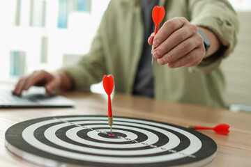 Man with dart aiming at dartboard while working on laptop indoors, closeup