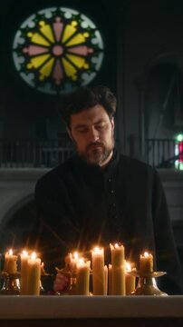 Vertical of middle-aged Caucasian clergyman in black soutane putting burning candle on altar while praying under candlelight in Catholic church with round stained glass window behind