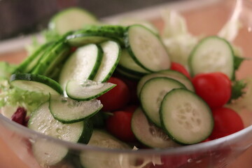 Fresh salad preparation with cucumbers, cherry tomatoes, and greens in a clear bowl on a kitchen countertop
