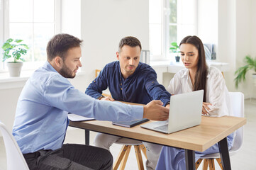 Young family couple sitting at desk with realtor, looking at laptop screen, choosing new house. Husband and wife exploring real estate options for purchase or rent with professional real estate agent.